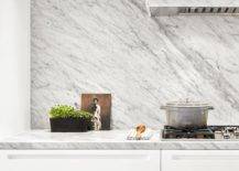 A marble clad hood is fixed to a marble slab backsplash over a gas integrated cooktop fixed to a gray marble countertop finishing white cabinets.