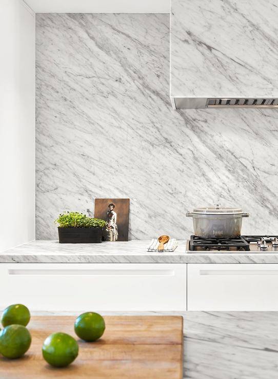 A marble clad hood is fixed to a marble slab backsplash over a gas integrated cooktop fixed to a gray marble countertop finishing white cabinets.