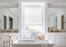 Master bathroom features a freestanding bathtub with nickel tub filler in front of a marble window seat flanked by French washstands topped with marble, lit by an Amber French chandelier hung from a tray ceiling and a gold accent table.