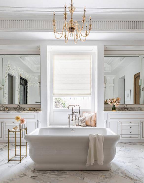 Master bathroom features a freestanding bathtub with nickel tub filler in front of a marble window seat flanked by French washstands topped with marble, lit by an Amber French chandelier hung from a tray ceiling and a gold accent table.