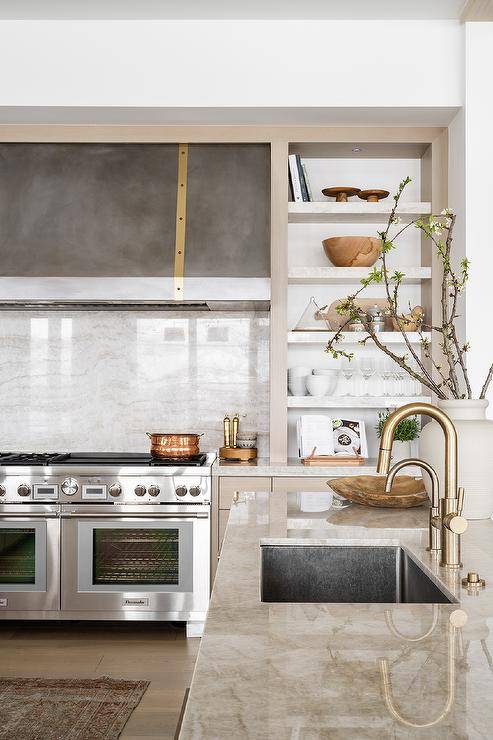 A steel and brass range hood is mounted to a beige marble slab cooktop backsplash over a Thermador dual range and between floating shelves.