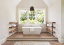 Industrial pipe and salvaged wood shelves create a unique feature flanking a rectangular freestanding tub completed with a brass faucet under a window and vaulted ceiling displaying a black hanging drum pendant. A step up feature adds to an appealing surface for the freestanding tub with a beige runner in front of the step.