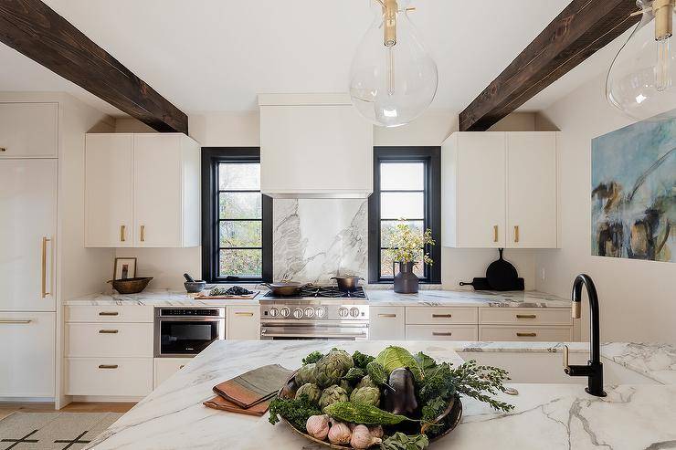 Rustic wood beams accent a white kitchen boasting a white range hood fixed between black framed windows to a marble backsplash over a stainless steel oven range. The range sits between white cabinets complemented with thick brass pulls.
