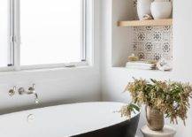 Cottage bathroom design features a modern black oval bathtub with a nickel wall mount tub filler, ivory and black mosaic tiles on the back of bathtub shelves and a white accent table.