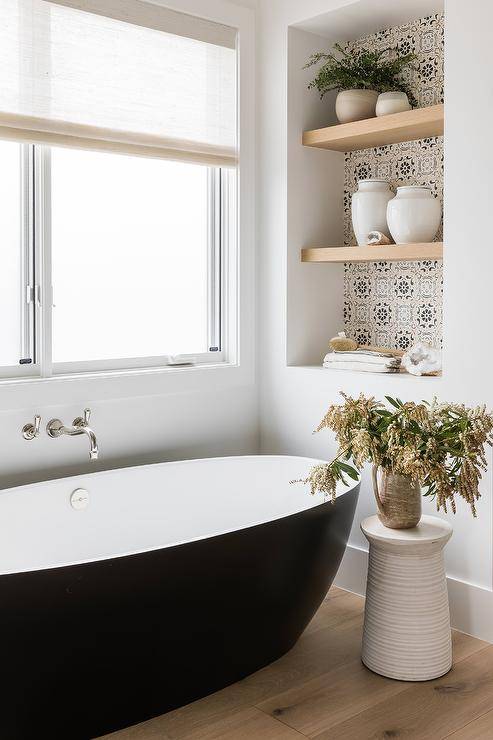 Cottage bathroom design features a modern black oval bathtub with a nickel wall mount tub filler, ivory and black mosaic tiles on the back of bathtub shelves and a white accent table.