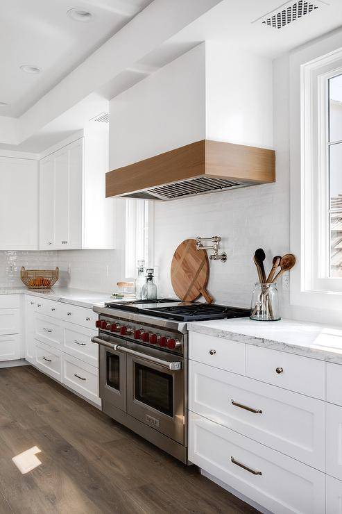 A white wooden hood is accented with beige wood trim placed above a white glazed tiled backsplash and a Wolf dual range.