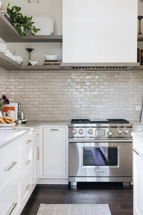 White kitchen features taupe glazed brick backsplash tiles with gray floating shelves, a wolf range under a white hood and white cabinets with nickel pulls.