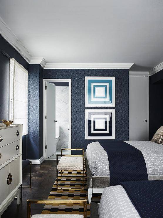 Beautifully styled shared bedroom features a polished brass bench placed on a yellow, black, and gray rug at the foot of gray velvet beds dressed in gray and blue bedding. Blue geometric art is stacked against a blue wallpapered wall beside an en suite bathroom door.