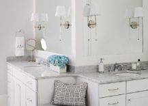 A built-in corner bench fitted with a single drawer adorned with a polished nickel pull topped with a gray cushion is flanked by his and her washstands topped with carrera marble placed under Camille Long Sconces mounted on thick, white framed vanity mirrors.