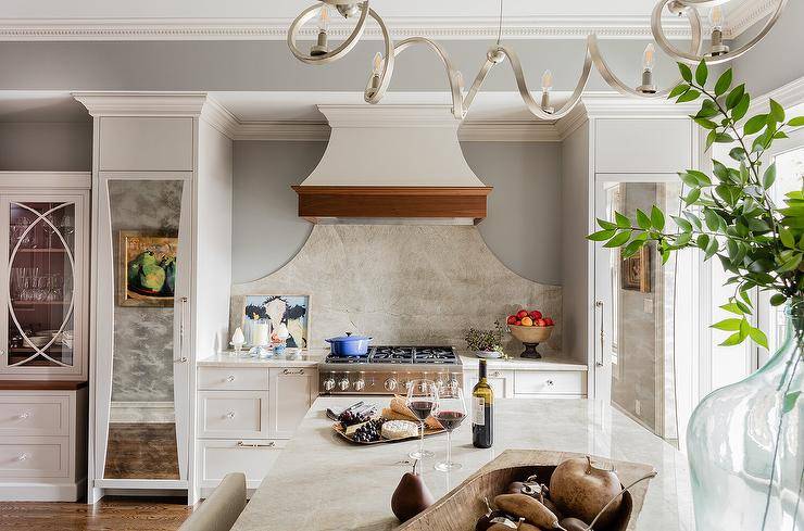 A gorgeous white French range hood with brown wood trim is mounted in a cooking alcove over a curved marble cooktop backsplash and an oven range flanked by white cabinets.