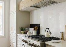 A white range hood with reclaimed wood trim is fixed against a marble-look quartz backsplash over a stainless steel oven range flanked by light gray cabinets donning brass hardware and a white quartz countertop.