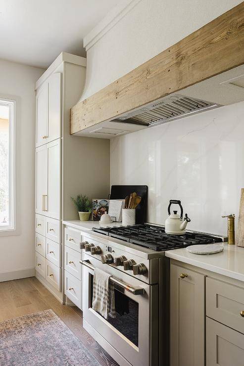A white range hood with reclaimed wood trim is fixed against a marble-look quartz backsplash over a stainless steel oven range flanked by light gray cabinets donning brass hardware and a white quartz countertop.