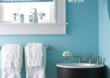 White and turquoise powder room features a round espresso corner vanity framed by bright turquoise blue walls and illuminated by a tiered capiz chandelier situated beside a small window dressed in a blue and white striped roman shade.