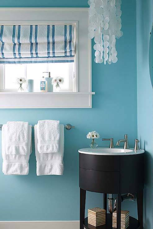 White and turquoise powder room features a round espresso corner vanity framed by bright turquoise blue walls and illuminated by a tiered capiz chandelier situated beside a small window dressed in a blue and white striped roman shade.