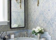 Gorgeous blue and gray bathroom with gray corner bow front bathroom vanity paired with blue stone countertop and black rectangular bathroom mirror, Blue and gray flocked wallpaper in bathroom and antique brass French sconces.