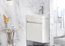 A modern corner vanity finished in all while flanked by a marble-styled wall and a sleek metal table.