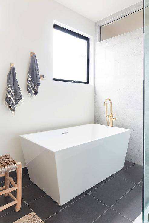 Bathroom features a modern white freestanding rectangular bathtub with polished brass gooseneck faucet tub filler on a black tiled floor.
