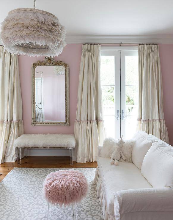 White and pink French bedroom features silk ruffled curtains, a baroque mirror over a white faux fur bench and a white ruffled sofa with pink faux fur stool atop a gray animal print rug illuminated by a feather chandelier.