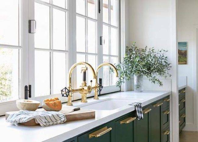 Stunning green kitchen cabinets are adorned with brass hardware complementing polished brass gooseneck faucets mounted side-by-side over an undermount sink and in front of a row of windows.