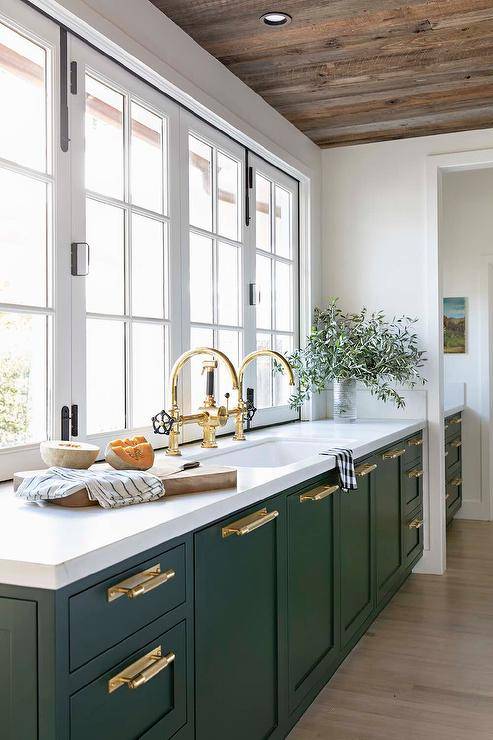 Stunning green kitchen cabinets are adorned with brass hardware complementing polished brass gooseneck faucets mounted side-by-side over an undermount sink and in front of a row of windows.