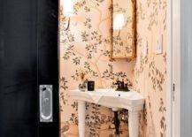 Clad in pink chinoiserie wallpaper, this beautiful bathroom features a corner Parisian pedestal sink accented with an oil rubbed bronze faucet kit located under a corner mounted gold ornate mirror. The sink sits on marble hex floor tiles that lead to a black pocket door.