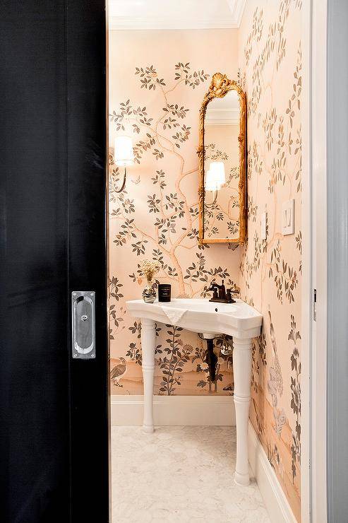 Clad in pink chinoiserie wallpaper, this beautiful bathroom features a corner Parisian pedestal sink accented with an oil rubbed bronze faucet kit located under a corner mounted gold ornate mirror. The sink sits on marble hex floor tiles that lead to a black pocket door.