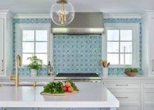Morgan Harrison Home - A stainless steel range hood is mounted against blue mosaic backsplash tiles and is positioned between windows and over a stainless steel range flanked by white drawers donning brass pulls and a white quartz countertop.
