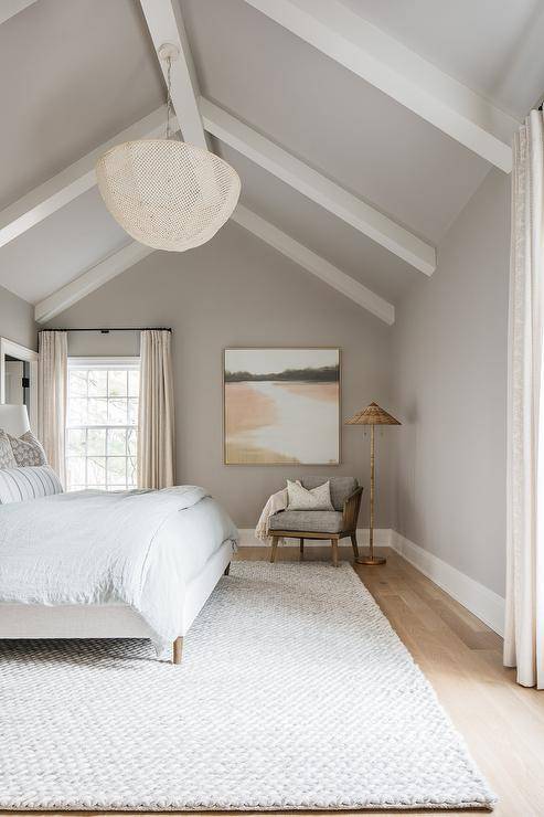 A white basket chandelier hangs from a gray vaulted ceiling accented with white wood beams over a white upholstered bed placed on white rug, as a gray accent chair is placed beneath an art piece hung from a gray wall beside a rattan floor lamp.