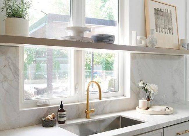A long floating shelf is mounted in front of a window partially framed by a marble slab backsplash and located over light gray cabinets donning brass hardware and a marble countertop holding a stainless steel sink with a brushed gold gooseneck faucet.