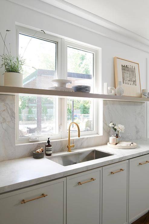 A long floating shelf is mounted in front of a window partially framed by a marble slab backsplash and located over light gray cabinets donning brass hardware and a marble countertop holding a stainless steel sink with a brushed gold gooseneck faucet.