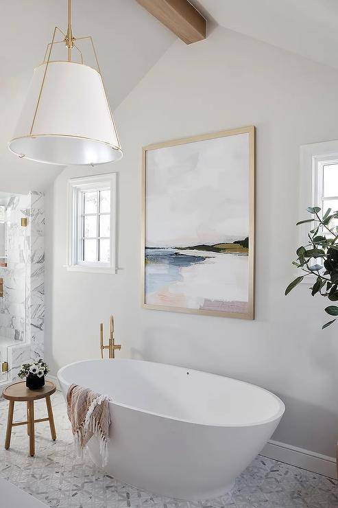 Bathroom features an oval bathtub with a brass floor mount tub filler on gray parquet marble tiles and a round accent table.