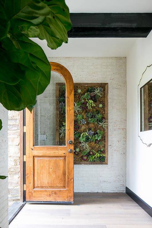 An arched front door with a single glass panel opens to light gray wash wood floors leading to a white framed mirror hung from a white wall. A succulents garden hangs from an exposed brick white wall beneath a ceiling accented with black wood ceiling beams.