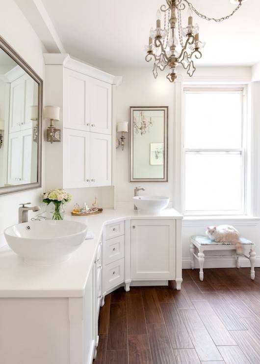 White bathroom features curved cabinets topped with his and her bowl sinks paired with off-set faucets and beveled mirrors flanking corner center console cabinet accented with antiqued sconces illuminated by crystal chandelier.