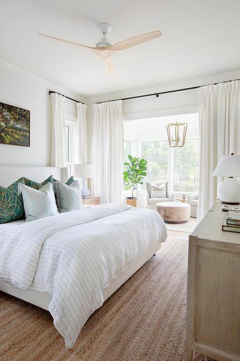 The cozy master bedroom living area boasts a round ottoman set in front of a dove gray wingback chair lit by a brass lantern hanging in front of the window, in front of a nook that has been rubbed with oil. It has white curtains hanging from bronze bars.