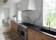 A white range hood is fixed against a honed marble slab backsplash between windows and over an aged brass swing arm pot filler and a stainless steel dual range. A black marble countertop accents brown cabinets fitted with a trough sink and a brass gooseneck faucet.