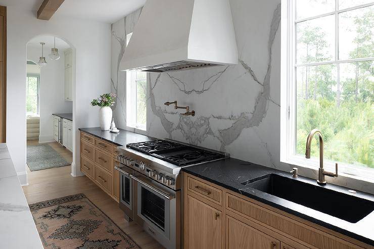 A white range hood is fixed against a honed marble slab backsplash between windows and over an aged brass swing arm pot filler and a stainless steel dual range. A black marble countertop accents brown cabinets fitted with a trough sink and a brass gooseneck faucet.