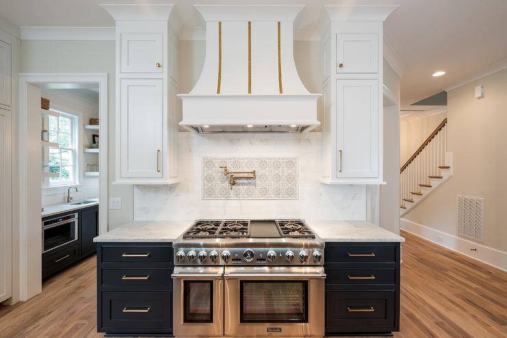 White and gold French range hood designed at a cooktop designed with mosaic accent tiles, an antique swivel arm pot filler and a thermador dual range.