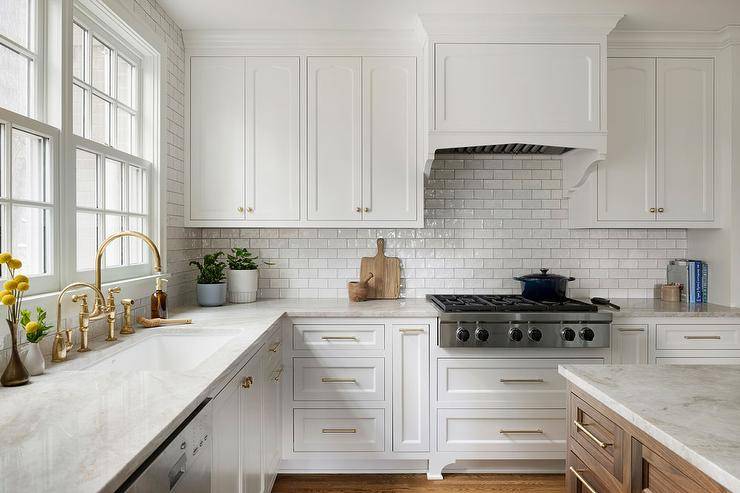 Kitchen features a white wooden range hood with white glazed backsplash tiles with light gray grout, white cabinets and an aged brass deck mount faucet.