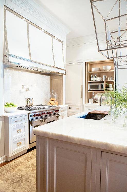 Kitchen features Darlana lanterns over a gray center island, a microwave cabinet with folding doors and a stove alcove with a white French range hood with stainless steel straps over a nickel swing arm pot filler and a Wolf dual range.