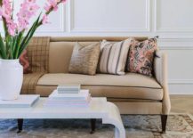Beige and pink living room features a white lacquer waterfall coffee table placed on a brown wool rug in front of a beige sofa finished with brass nailhead trim and pink pillows. White walls are accented with white millwork.