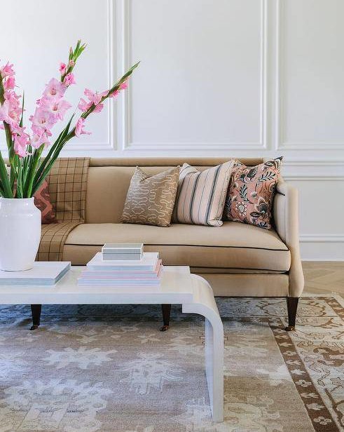 Beige and pink living room features a white lacquer waterfall coffee table placed on a brown wool rug in front of a beige sofa finished with brass nailhead trim and pink pillows. White walls are accented with white millwork.