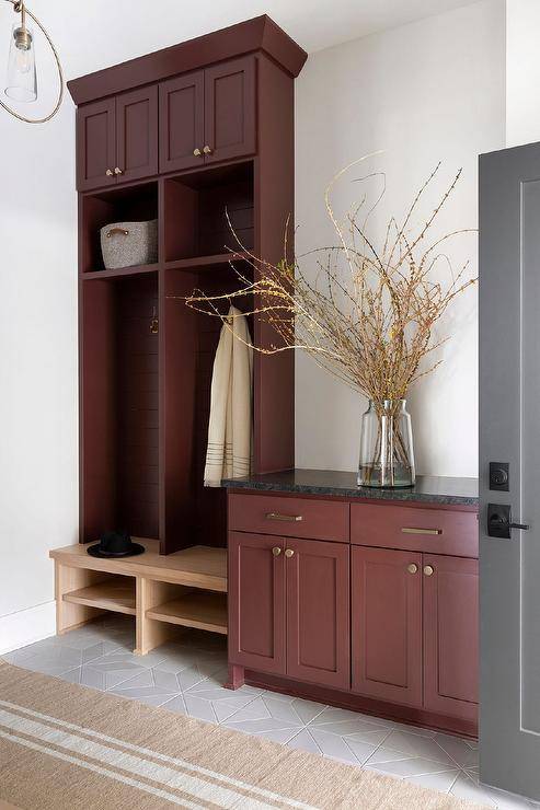 Burgundy red mudroom cabinets accented with aged brass hardware are topped with a black marble countertop and fixed beside burgundy red open lockers fitted with brass hooks mounted over a light stained wood bench. A french burlap runner covers gray feather style floor tiles.