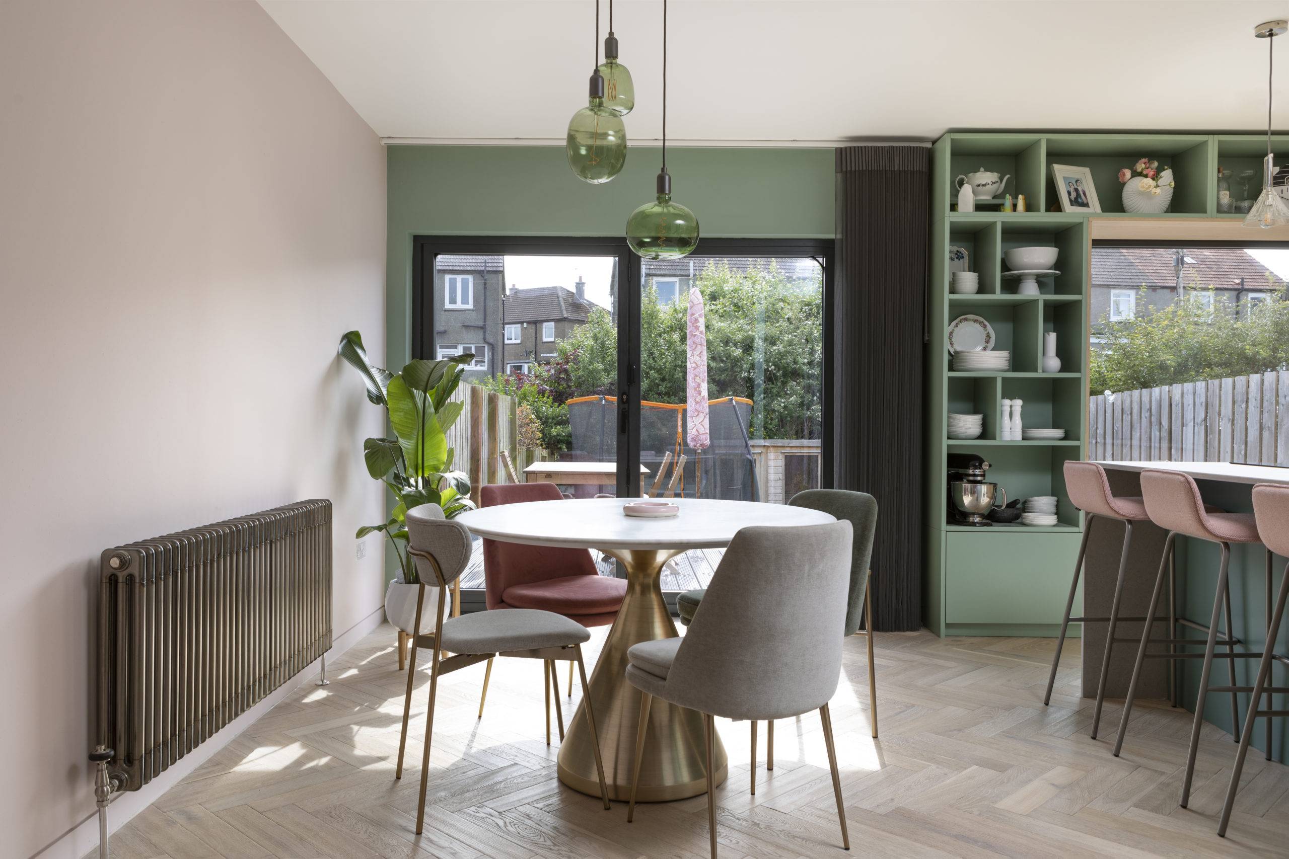 oak herringbone floors in kitchen with small circular dinning table and gold accented chairs, large sliding doors to exterior, green bespoke floor to ceiling shelving