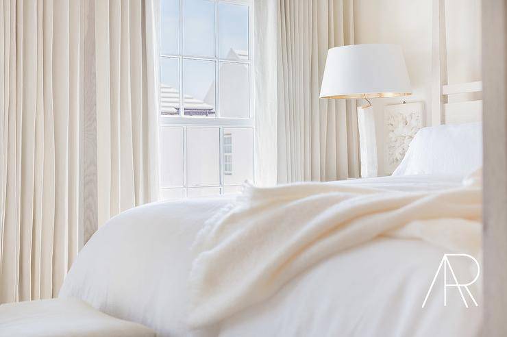 A restful contemporary white and ivory bedroom features white and gold nightstands next to a cream wood four-poster bed accented by white bedding with a cream cashmere blanket. illuminated by a white lamp with a lampshade. The windows are covered with gorgeous cream curtain panels.