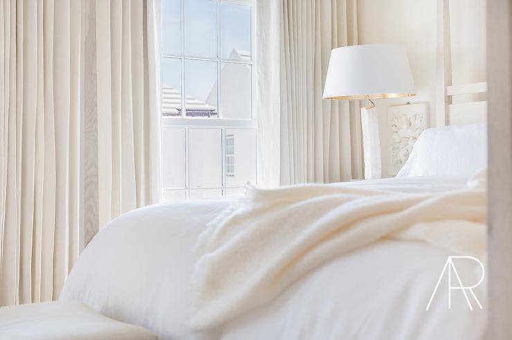 Restful, contemporary, white and ivory bedroom is lit by a white lamp with a white and gold lamp shade placed on a nightstand abreast a surf wood 4 poster bed accented with white sheets draped in a surf cashmere blanket. A window is covered with gorgeous surf drapery panels.