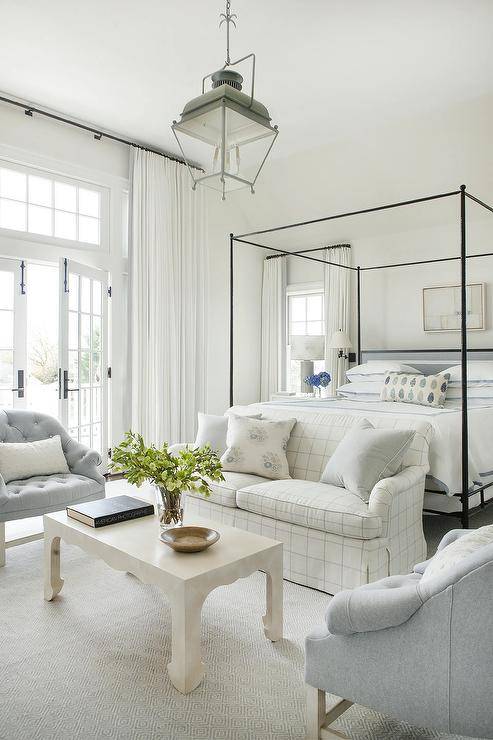 In front of a black canopy bed, a white and gray check roll arm sofa sits on a gray rug facing a cream coffee table flanked by gray tufted accent chairs lit by a carriage lantern.