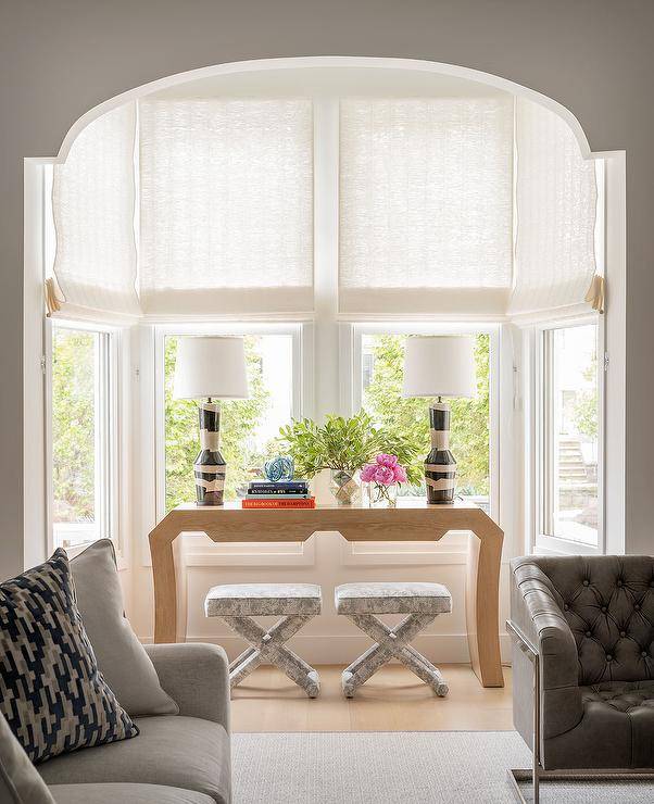 In an arched bay window nook, a blond oak waterfall console table is placed over gray x-stools and topped with black and white lamps lighting windows dressed in white roman shades.