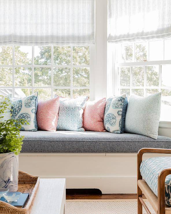 Light gray striped roman shades hang from bay windows over a white built-in bench topped with a blue cushion and pink and blue pillows.