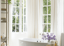 Bathroom features a bay window with oval bathtub, a round nickel and glass accent table and cream curtains.