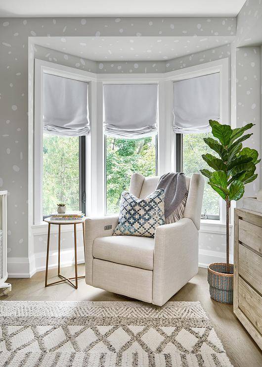 Contemporary nursery with a bay window furnished with an ivory wingback rocker and a fiddle leaf fig plant. Gray roman shades adorn the bay windows surrounded by white and gray wallpaper walls.